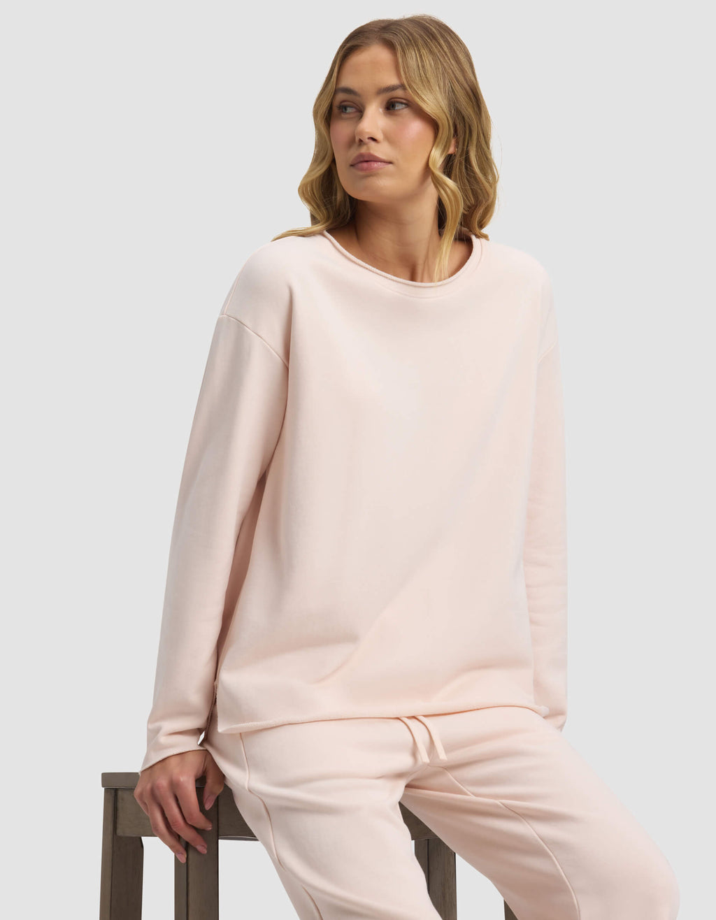 A woman with wavy blonde hair sits on a wooden stool, wearing the Cozy Earth Women's Chelsea Pullover in light pink. She gazes to her left with a neutral expression against a plain light background.|Color:Petal