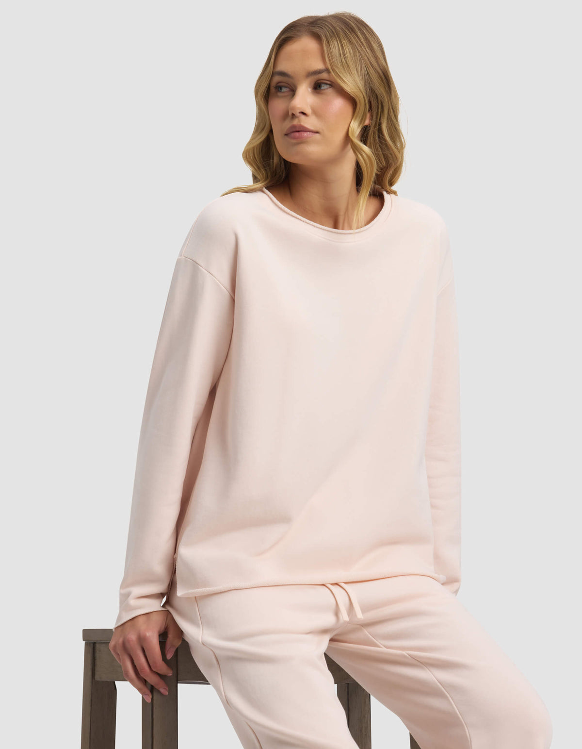 A woman with wavy blonde hair sits on a wooden stool, wearing the Cozy Earth Women's Chelsea Pullover in light pink. She gazes to her left with a neutral expression against a plain light background.|Color:Petal