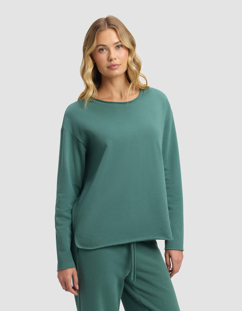 A woman with wavy blonde hair models the Cozy Earth Women's Chelsea Pullover and matching pants in green, standing against a plain light gray background.|Color:Sea Green