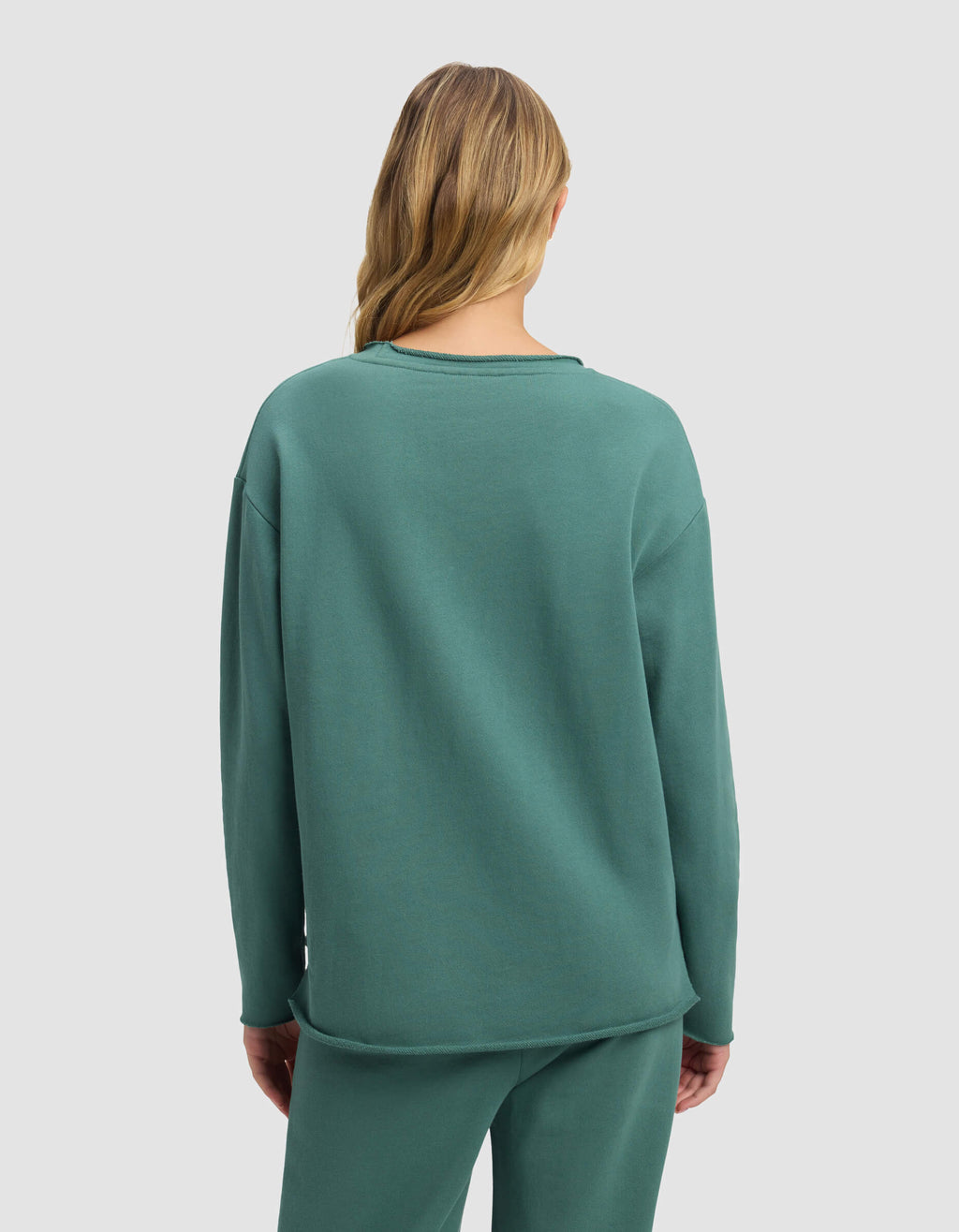A woman with long, wavy blonde hair is seen from behind wearing the Cozy Earth Women's Chelsea Pullover in teal, paired with matching pants, set against a plain light gray background.|Color:Sea Green