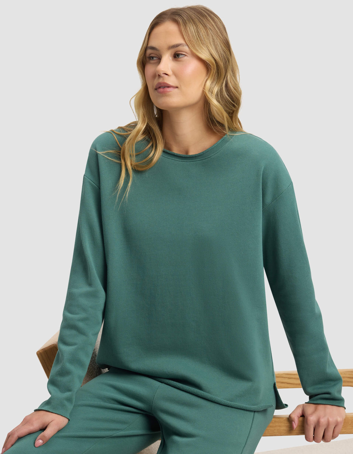 A woman with long blonde hair sits on a wooden bench wearing the Cozy Earth Women's Chelsea Pullover in teal, paired with matching pants, gazing slightly to the side against a plain background.|Color:Sea Green