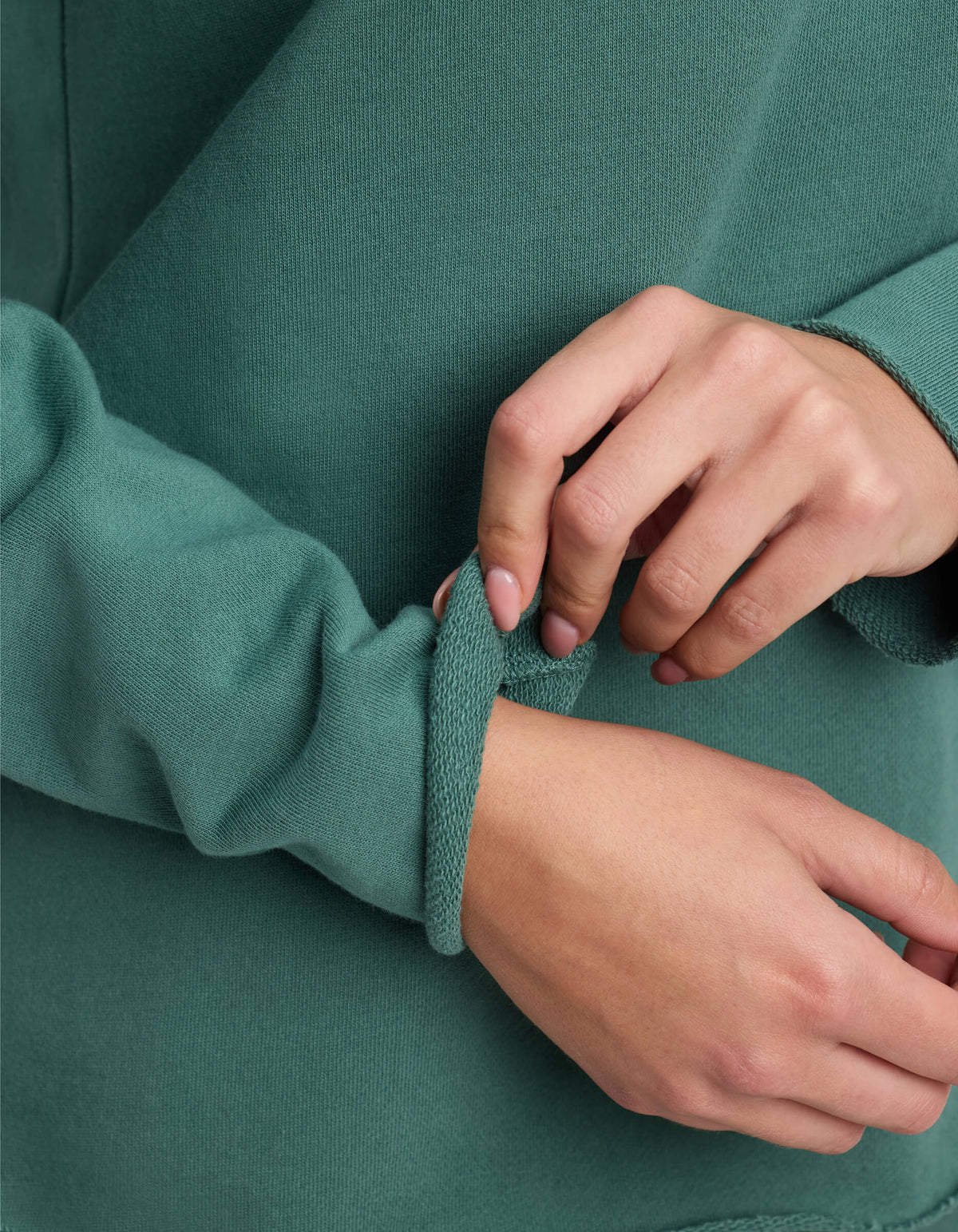 Close-up of a woman wearing the Cozy Earth Women's Chelsea Pullover in green, adjusting the cuff on her left sleeve with her right hand. The out-of-focus background highlights the hands and textured fabric.|Color:Sea Green