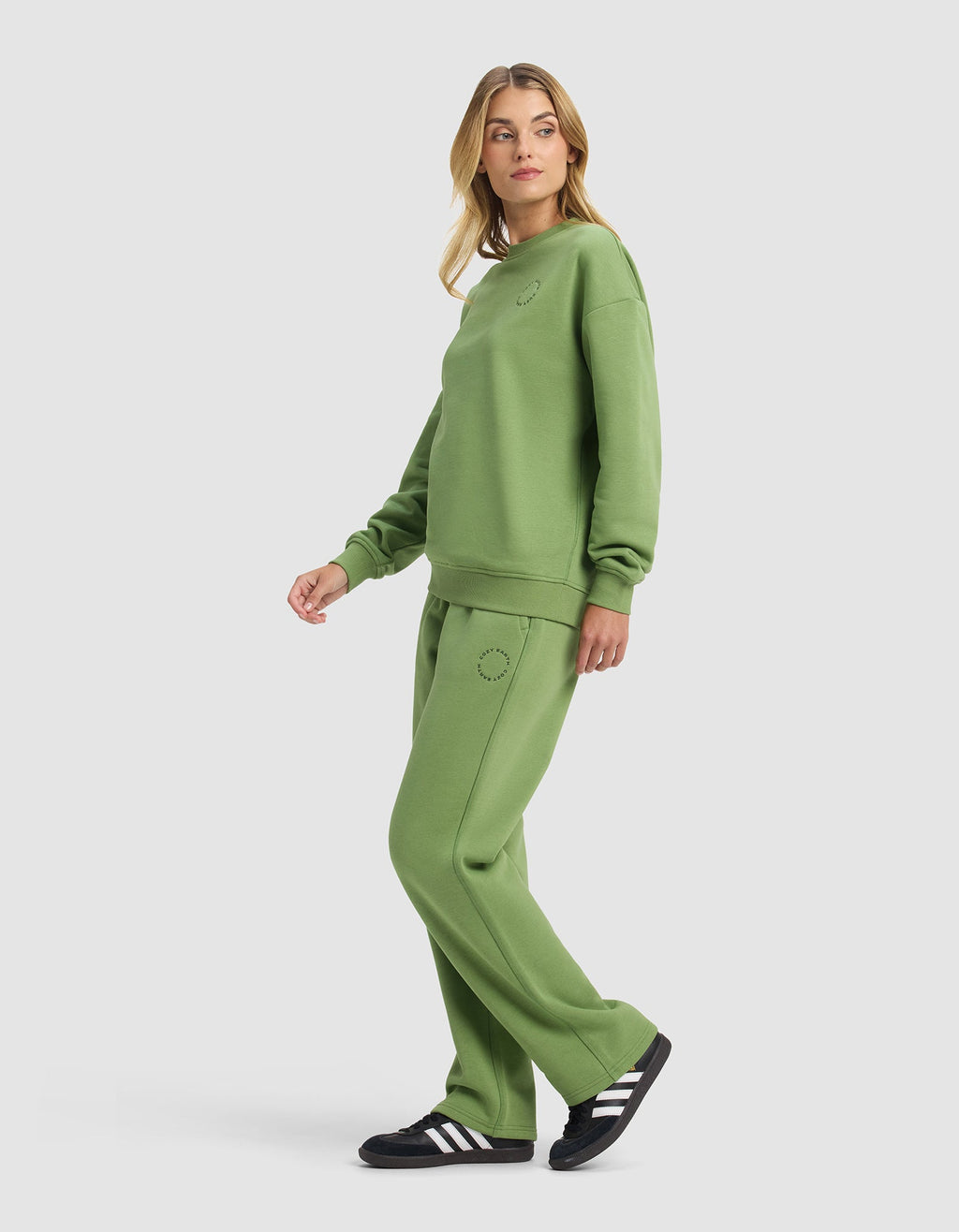 A woman stands sideways wearing the Cozy Earth Women's Cityscape Crewneck & Wide Leg Pant Set in green with black and white sneakers. The loose, casual outfit is shown against a plain white background as she looks over her shoulder.|Color:Clover
