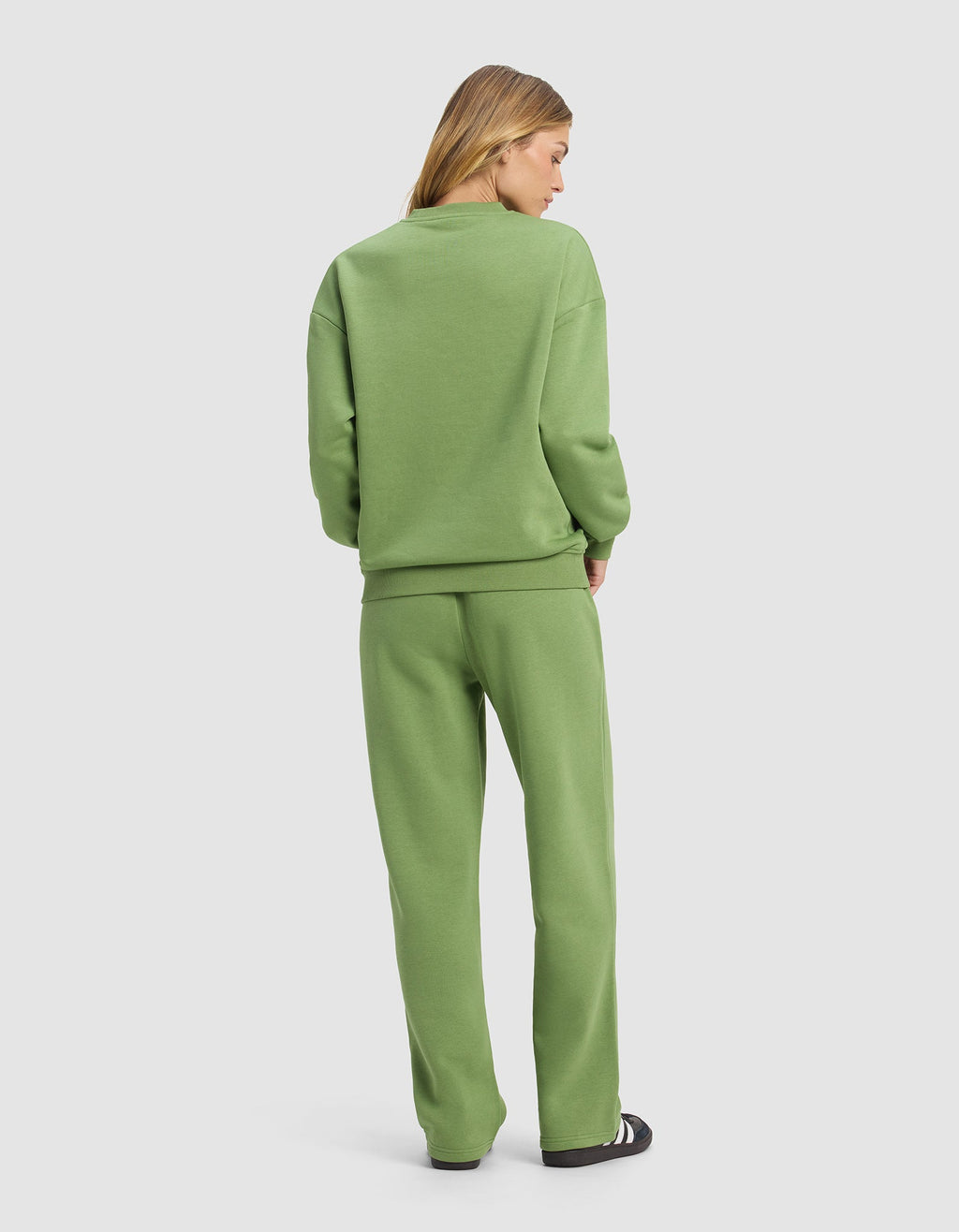 A woman faces away, wearing the Cozy Earth Women's Cityscape Crewneck & Wide Leg Pant Set in light green with black and white sneakers, standing against a plain light gray background.|Color:Clover