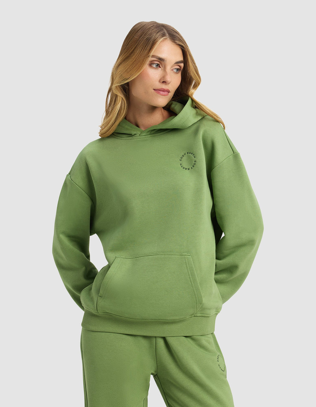 A woman with long blonde hair models the Cozy Earth Women's Cityscape Hoodie in green, paired with matching sweatpants, standing with hands in her hoodie pocket against a plain light gray background.|Color:Clover