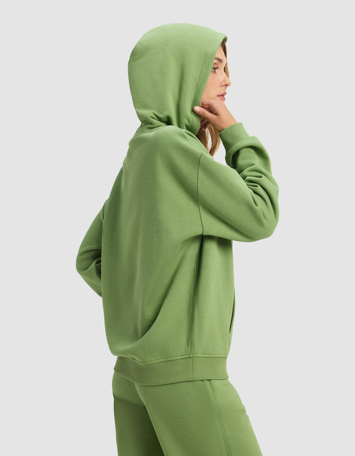 A woman in profile wears the Cozy Earth Women's Cityscape Hoodie and matching sweatpants in green, standing against a plain light background with her hood up and one hand touching her chin.|Color:Clover