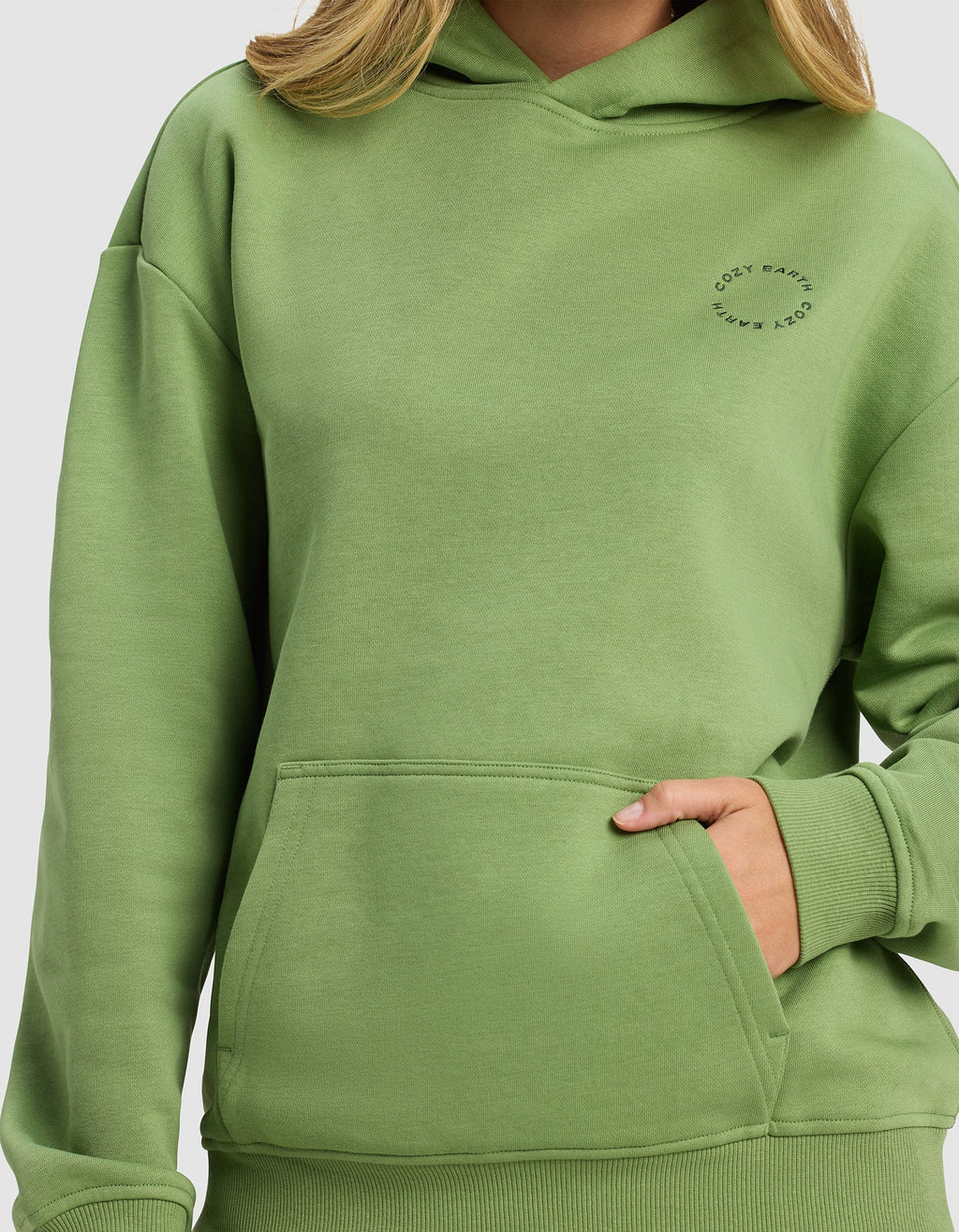 A woman wears the Cozy Earth Women's Cityscape Hoodie in light green, featuring a front pocket and small circular "COZY EARTH" text on the chest. Only her lower face, neck, and upper body are visible.|Color:Clover