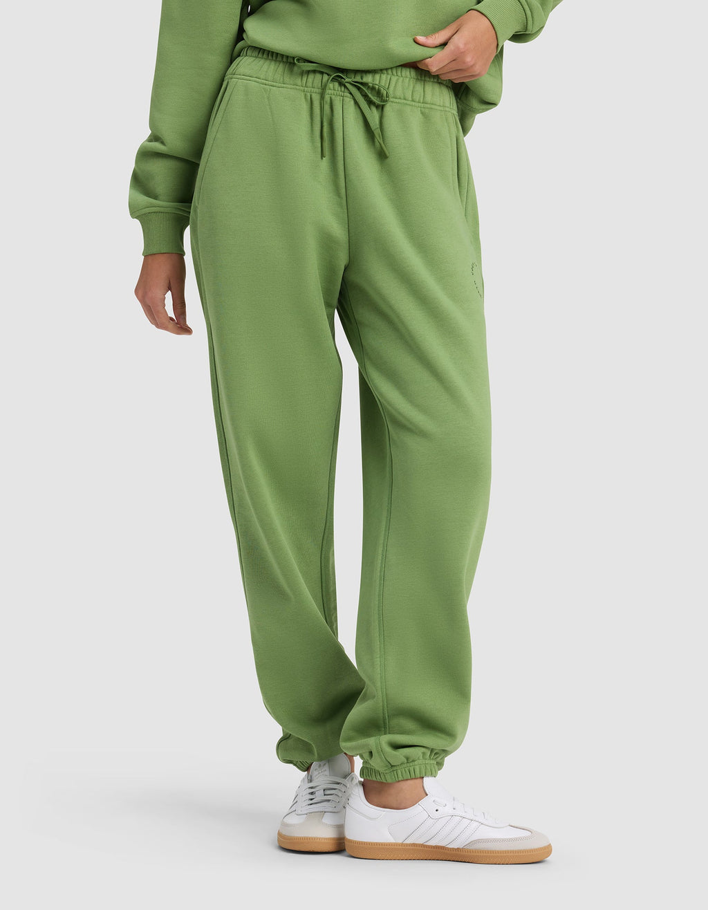 A person wears Cozy Earth's Women's Cityscape Sweatpant—a green pair with an elastic waistband and drawstring—styled with a matching sweatshirt and white sneakers, standing against a plain light background.|Color:Clover