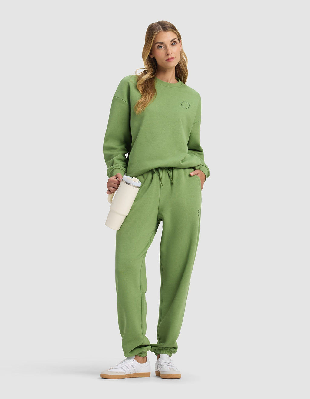 A woman models the Cozy Earth Women's Cityscape Sweatpant in green with a matching sweatshirt, white sneakers, and a cream water bottle, posing with one hand in her pocket against a plain light background.|Color:Clover