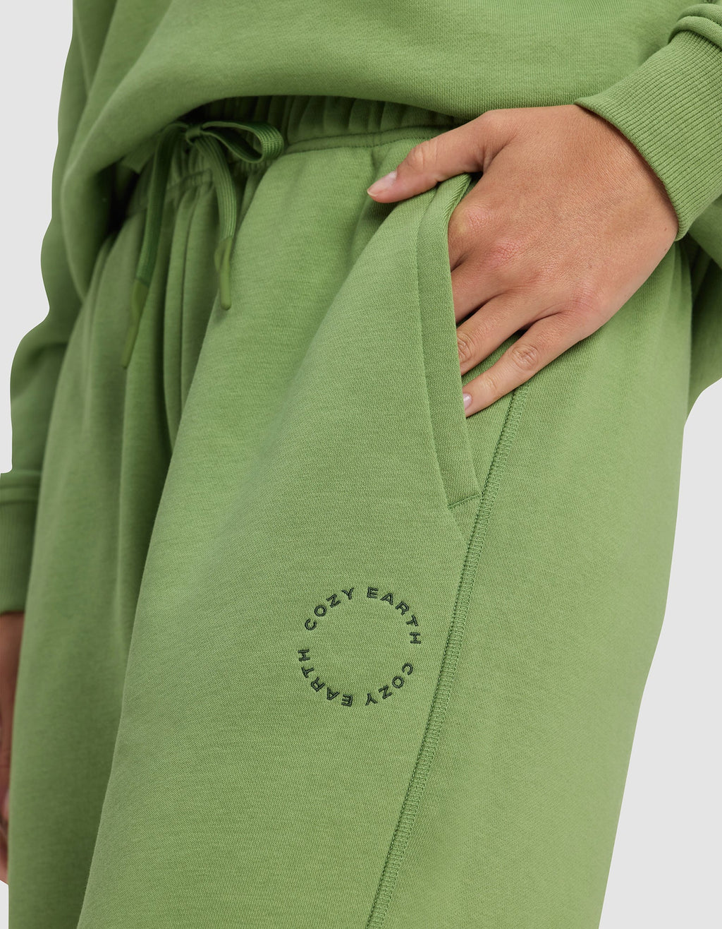 A person wears Cozy Earth's Women's Cityscape Sweatpant in light green with a matching sweatshirt, one hand in their pocket. "COZY EARTH" is printed in a circle near the sweatpant’s pocket.|Color:Clover