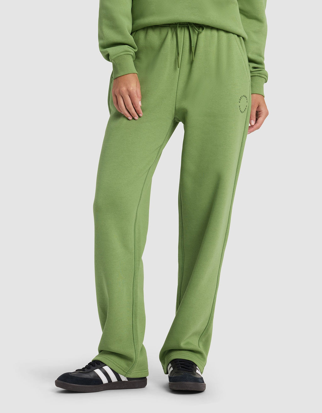 A woman wears Cozy Earth’s Women’s Cityscape Wide Leg Pant in green with a matching sweatshirt and black sneakers with white stripes, standing against a plain white background.|Color:Clover