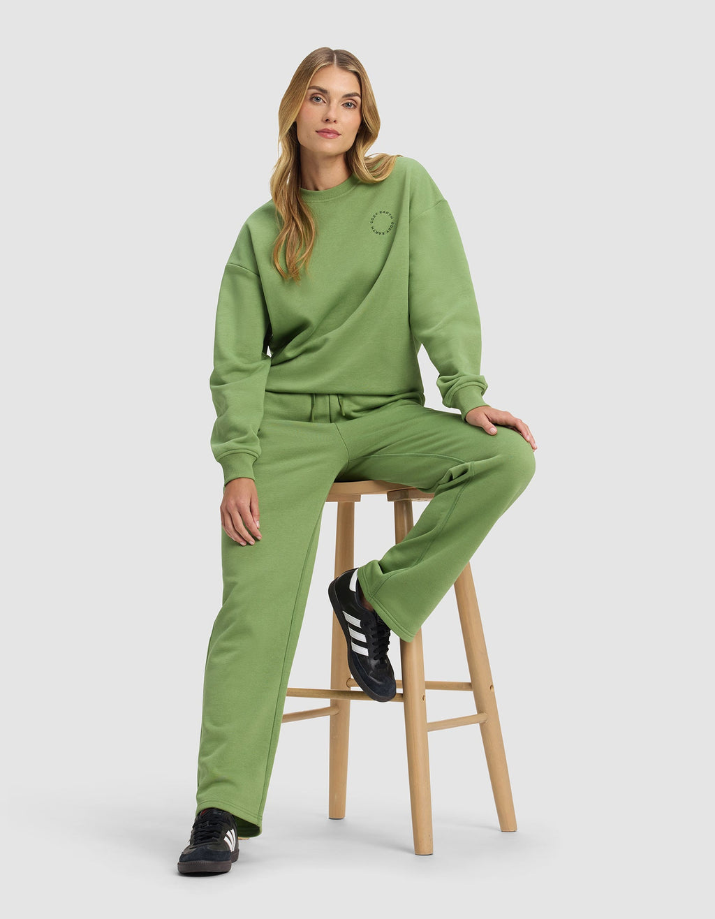A woman with long blond hair sits on a wooden stool, wearing Cozy Earth's Women's Cityscape Wide Leg Pant in green with a matching loose sweatshirt and black Adidas sneakers against a plain light gray background.|Color:Clover