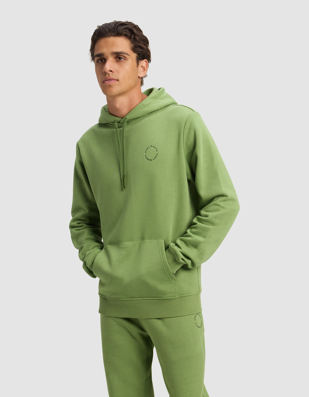 A man wears a Cozy Earth Men's Cityscape Hoodie and matching sweatpants in Clover, both featuring a small circular stitched design, with hands in the hoodie pocket against a plain light gray background.|Color:Clover