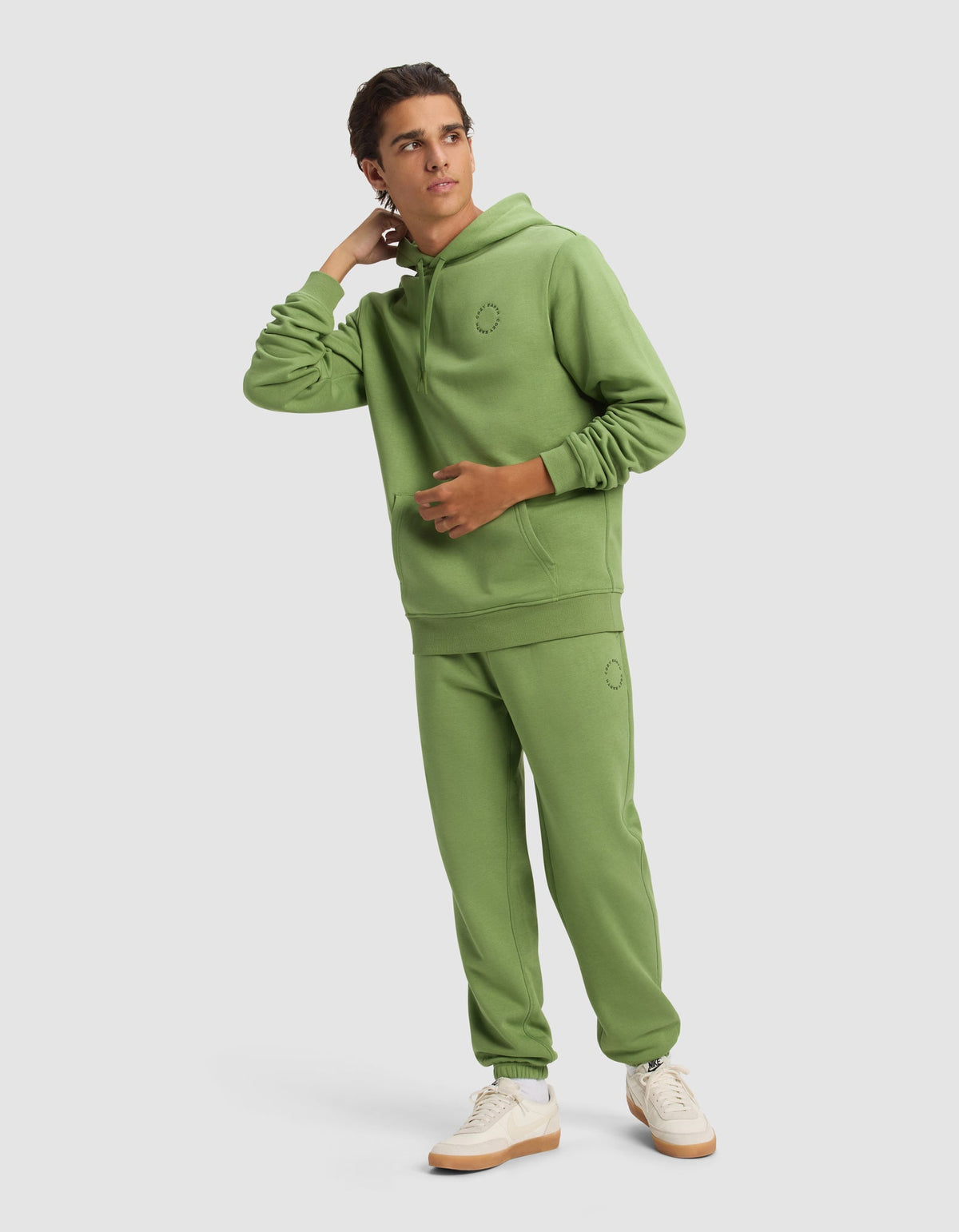 A young man in the Cozy Earth Men's Cityscape Hoodie and matching sweatpants with white sneakers stands against a plain background, looking to the side with one hand touching his neck.|Color:Clover