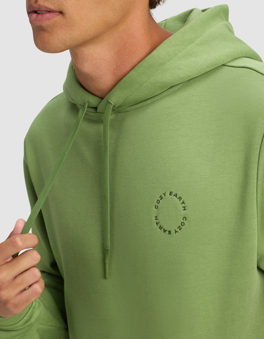 A man wears the Men's Cityscape Hoodie by Cozy Earth in light green, featuring a small circular logo on the chest. He holds a hoodie drawstring, with only his lower face visible.|Color:Clover