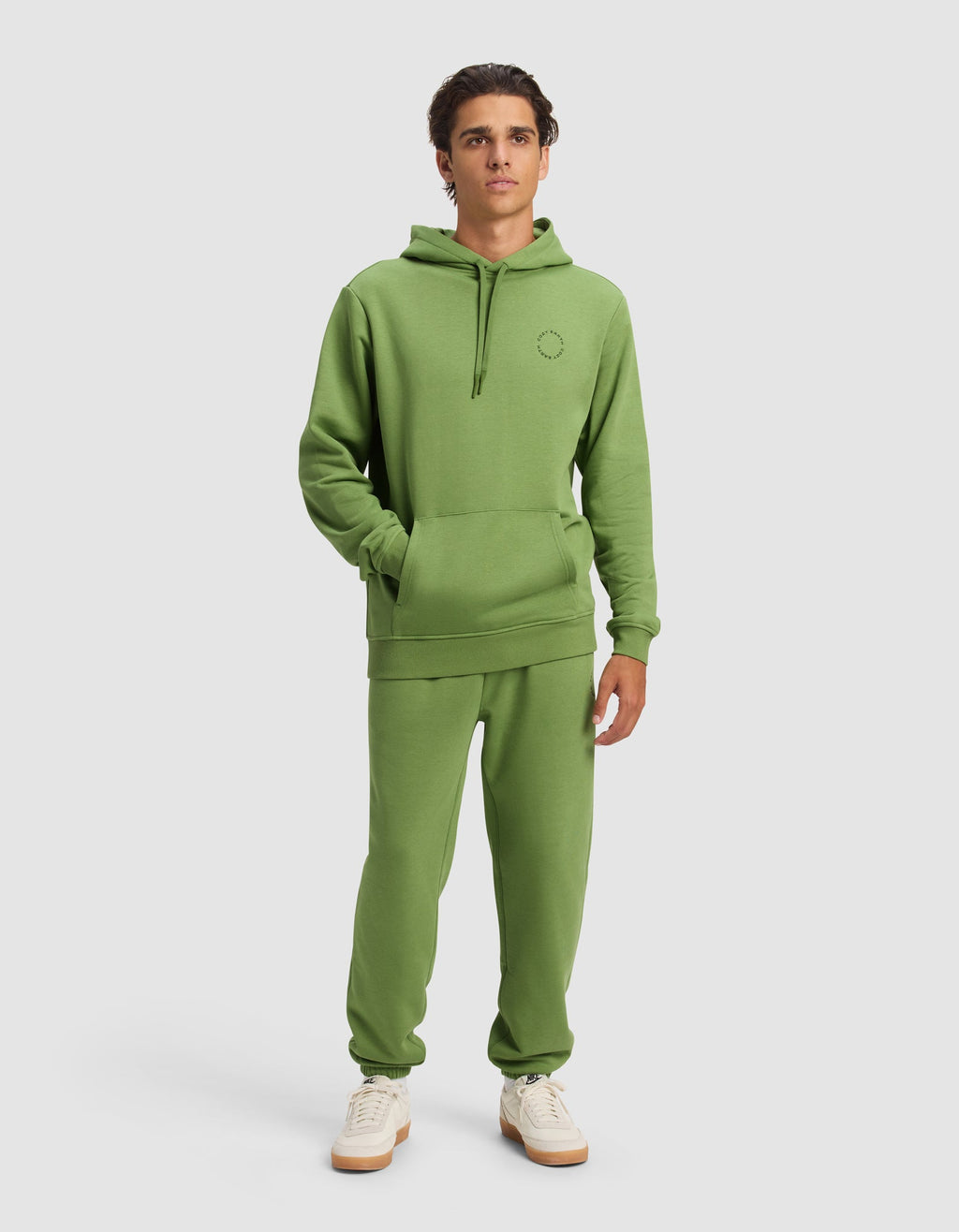 A young man wears the Cozy Earth Men's Cityscape Hoodie & Sweatpant Set with white sneakers, standing against a plain background with his hands in the hoodie pocket and a neutral expression.|Color:Clover