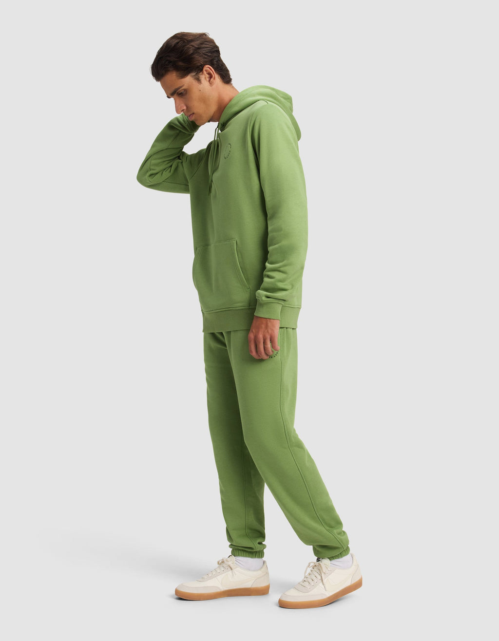 A man wears the Cozy Earth Men's Cityscape Hoodie & Sweatpant Set in light green, paired with white sneakers with tan soles, standing against a plain light background and touching the back of his neck.|Color:Clover