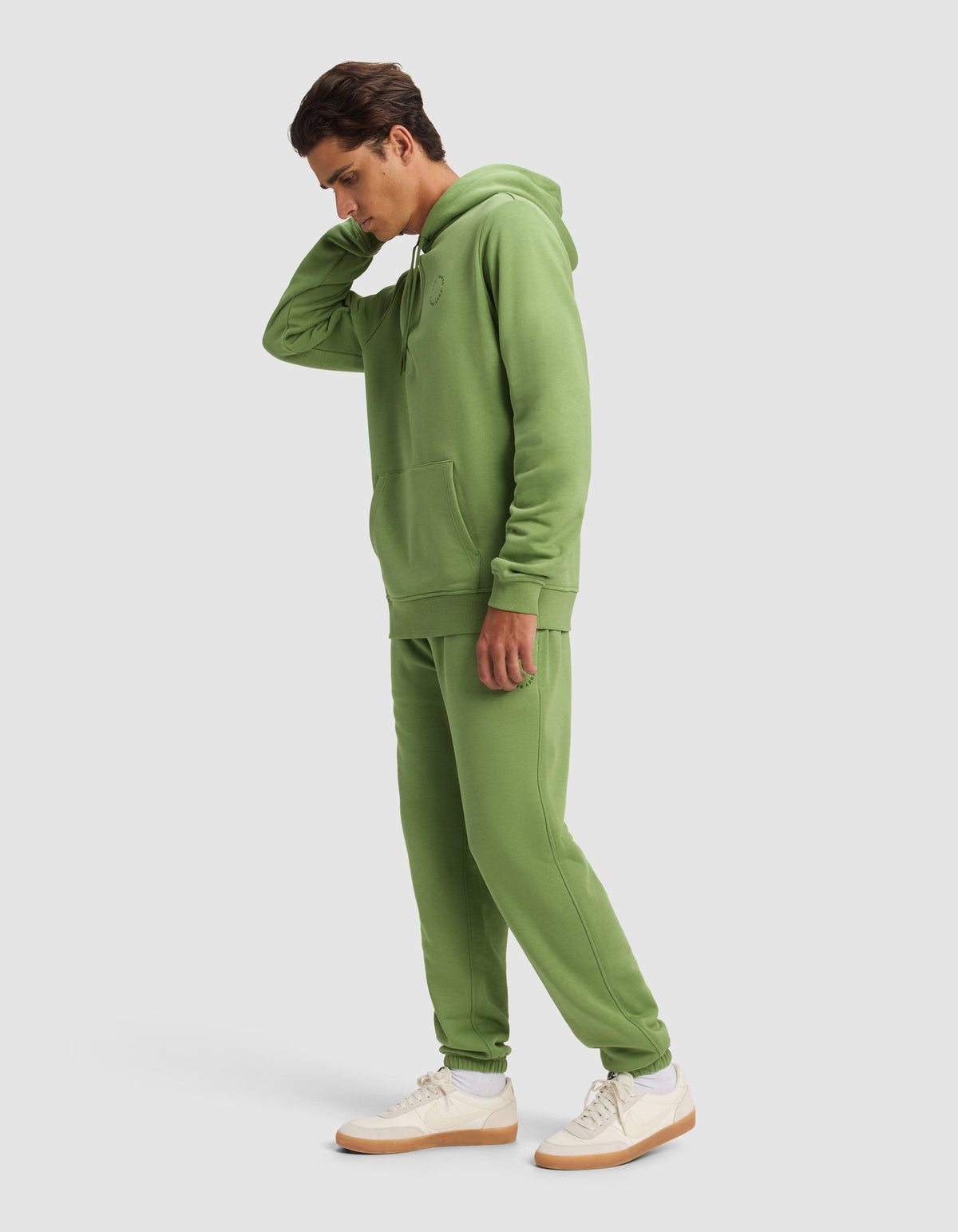 A man wears the Cozy Earth Men's Cityscape Hoodie & Sweatpant Set in light green, paired with white sneakers with tan soles, standing against a plain light background and touching the back of his neck.|Color:Clover
