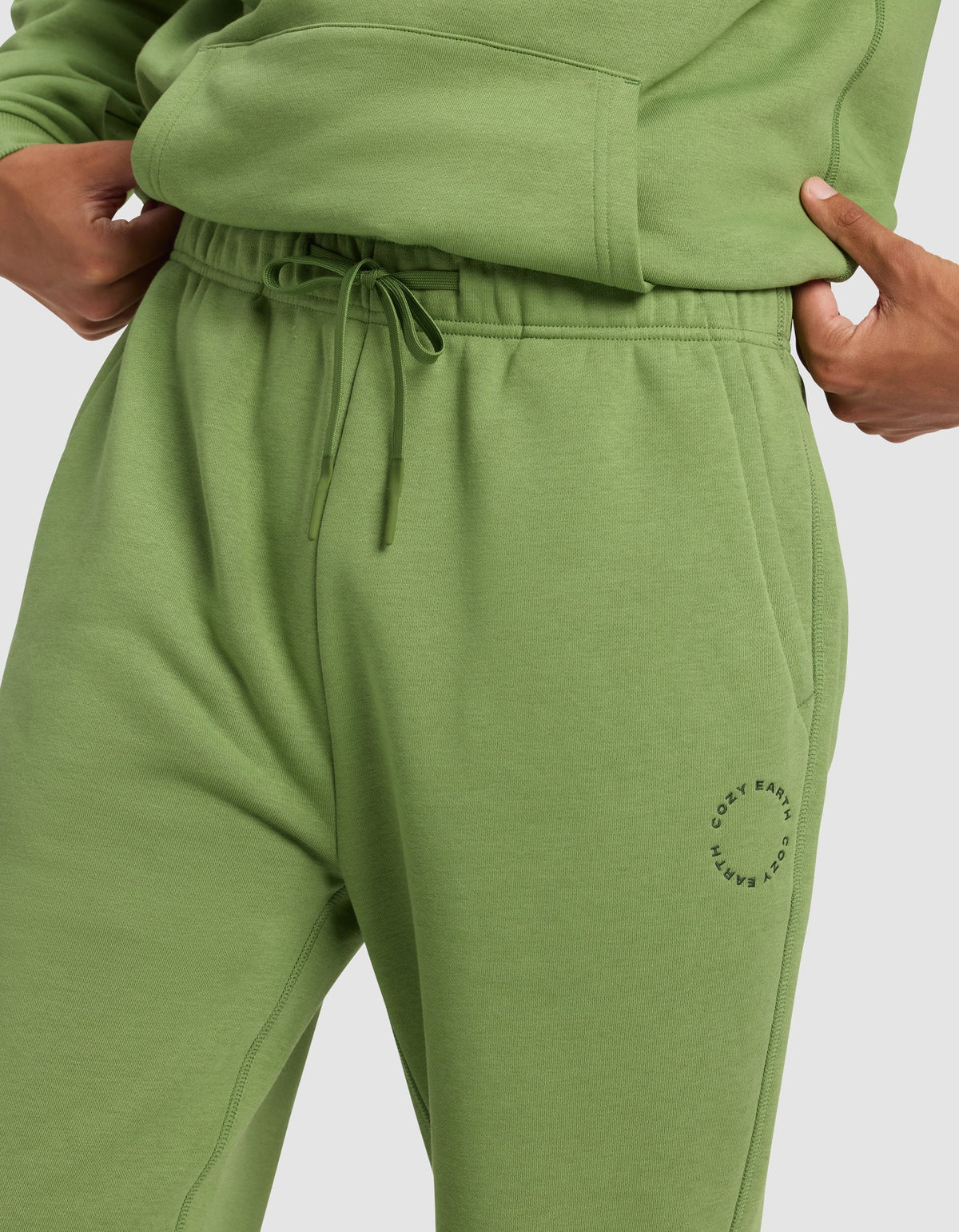 A person wears Cozy Earth Men's Cityscape Sweatpant in light green with an elastic drawstring waistband, a small circular logo by the left pocket, and a matching sweatshirt, one hand in the pocket.|Color:Clover