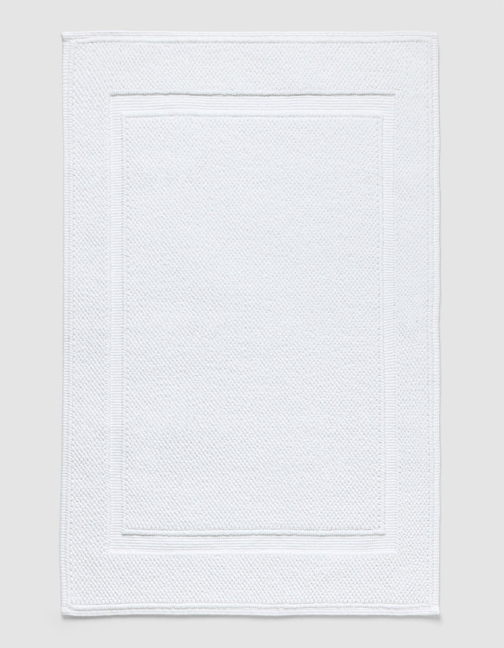 The Cozy Earth Classic Bath Mat in white features a textured surface with a subtle border pattern and is shown flat against a neutral background.|Color:White