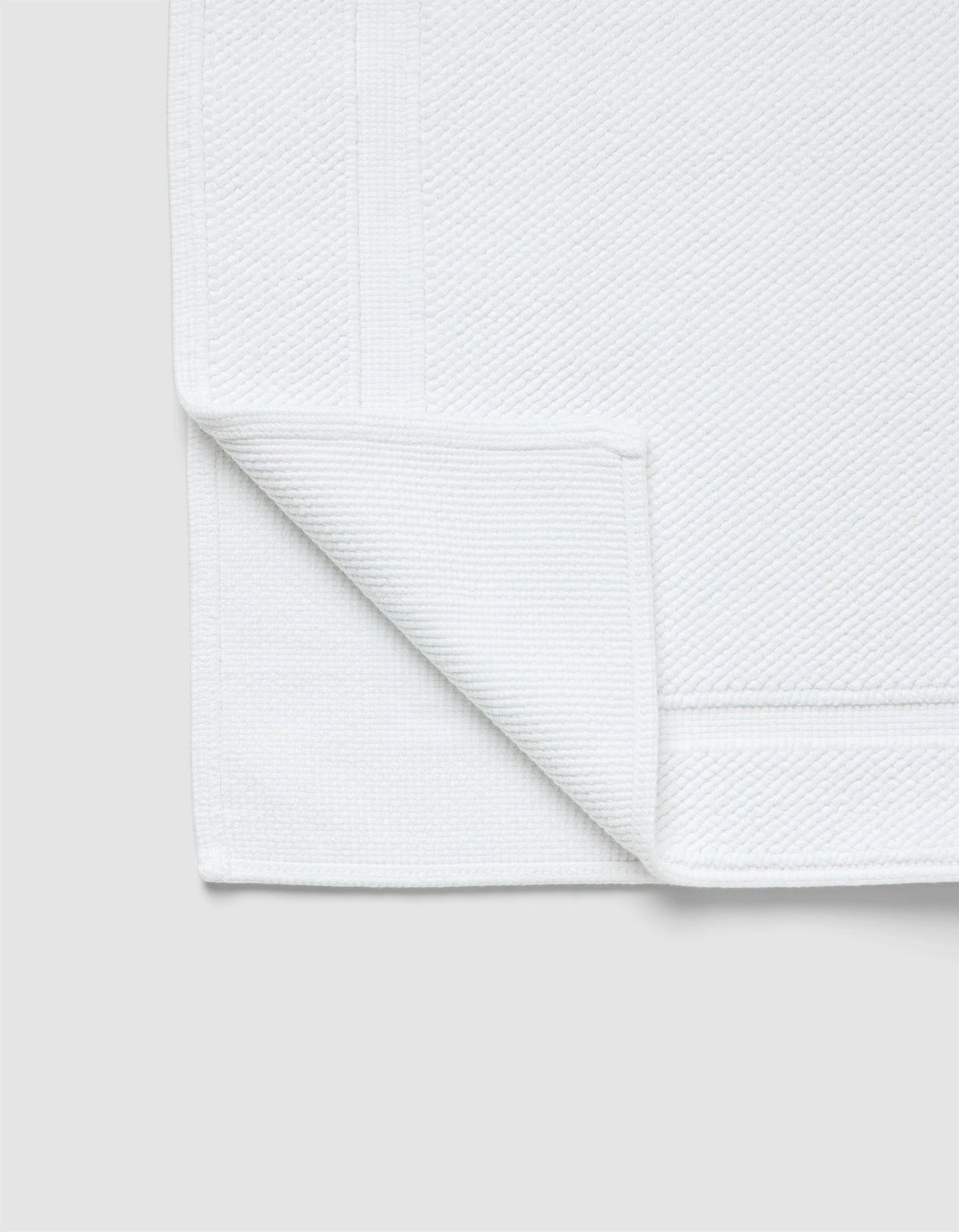 The Cozy Earth Classic Bath Mat, in white with a textured surface and simple border, is shown with one corner folded to reveal both sides against a light gray background.|Color:White