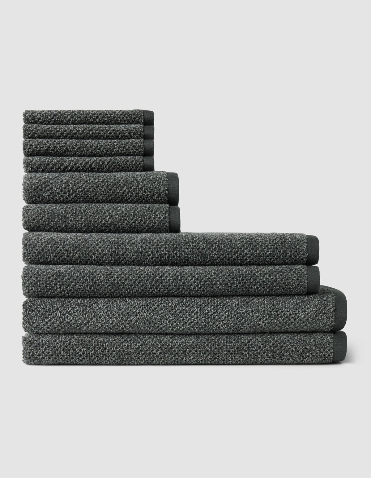 The Nantucket Bath Towel Set by Cozy Earth features a neatly stacked arrangement of dark gray towels in varying sizes, forming a stepped pattern against a plain light background.|Color:Graphite