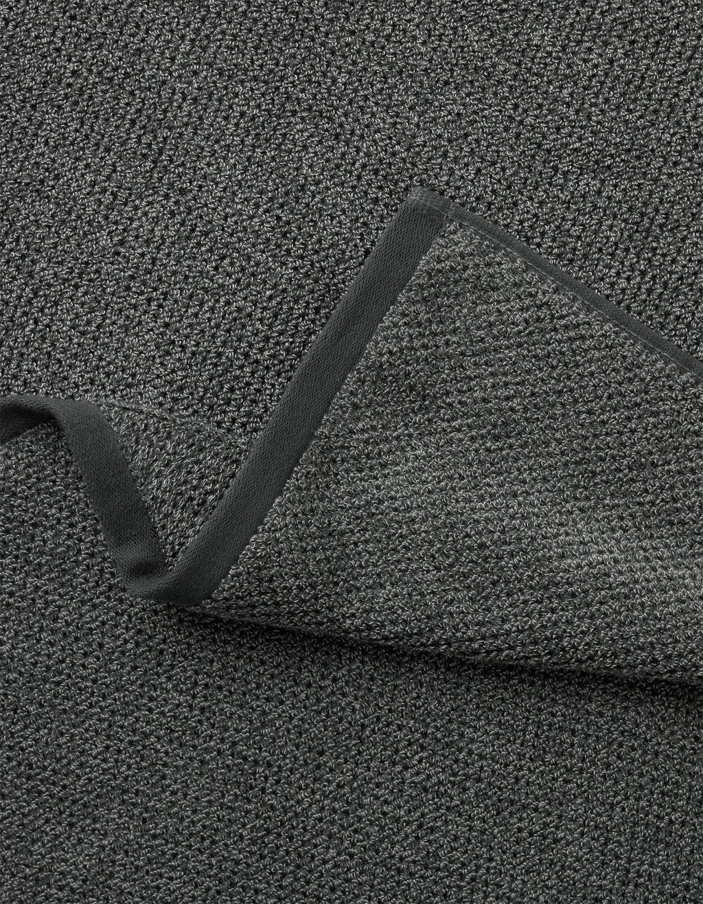 A close-up of the Cozy Earth Nantucket Bath Towel Set in dark gray, showing its textured fabric and black trim, with one corner folded to reveal its thickness.|Color:Graphite