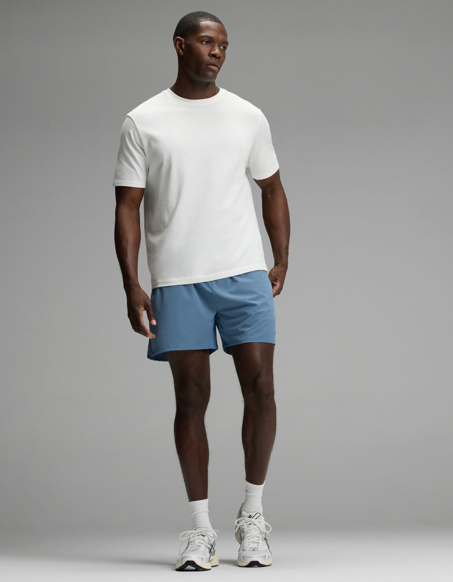 A man stands against a plain background wearing a white T-shirt, Cozy Earth Men’s Day & Night Short 5″ in blue, white socks, and white sneakers.|Color:Nordic Blue