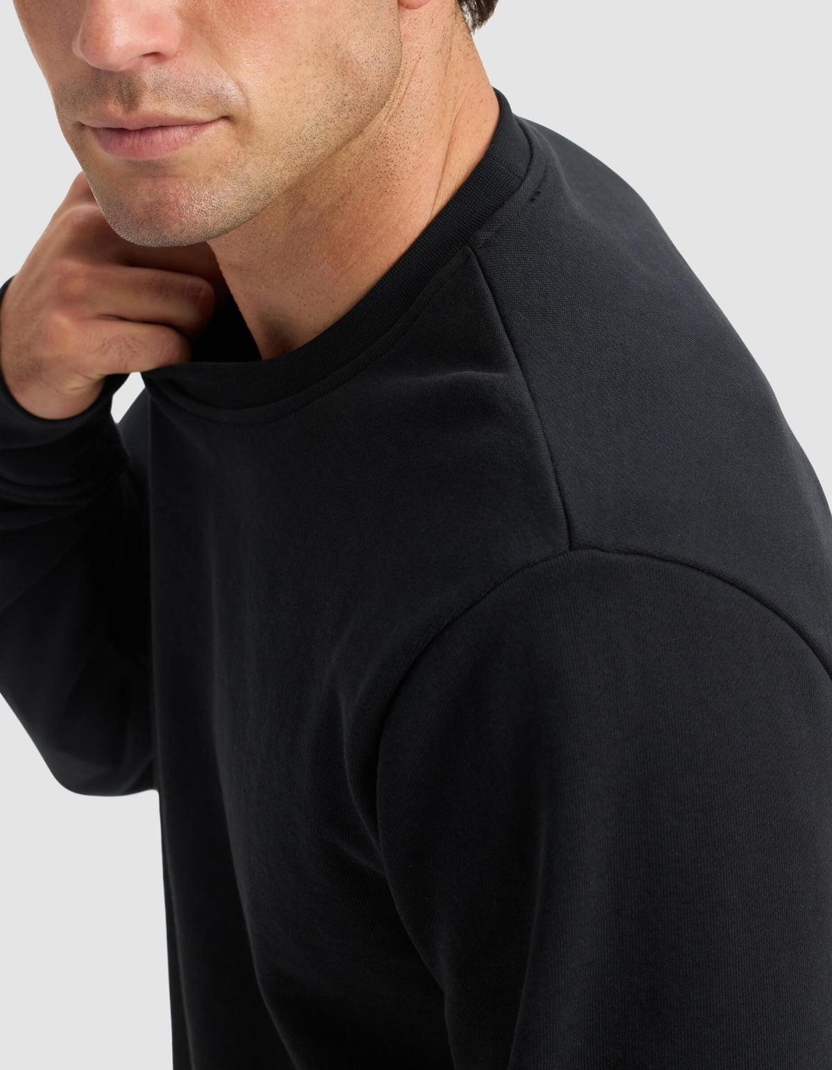 A man wearing the Cozy Earth Men's Dylan Crewneck is shown from the shoulders up, slightly turned to the side, with his hand touching the back of his neck against a light gray background.|Color:Jet Black