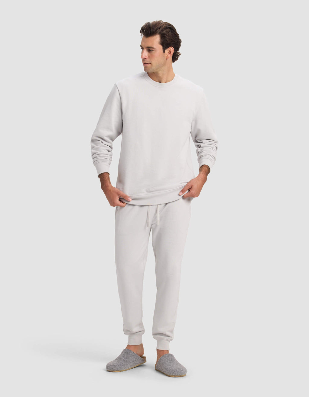A man wears the Cozy Earth Men's Dylan Jogger in light gray with matching joggers and slippers, standing against a plain, light background, looking to his left with hands near his waist.|Color:Dew