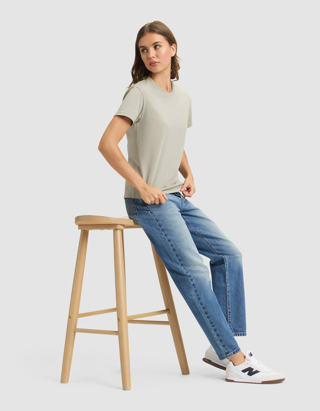 A woman wearing the Cozy Earth Women's Easy Tee in light green, blue jeans, and white sneakers sits sideways on a wooden stool against a plain light gray background. |Color:Palm