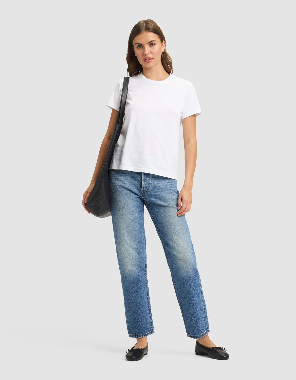 A woman stands against a plain background, wearing the Cozy Earth Women's Easy Tee in white, blue jeans, black flats, and carrying a black shoulder bag. |Color:White