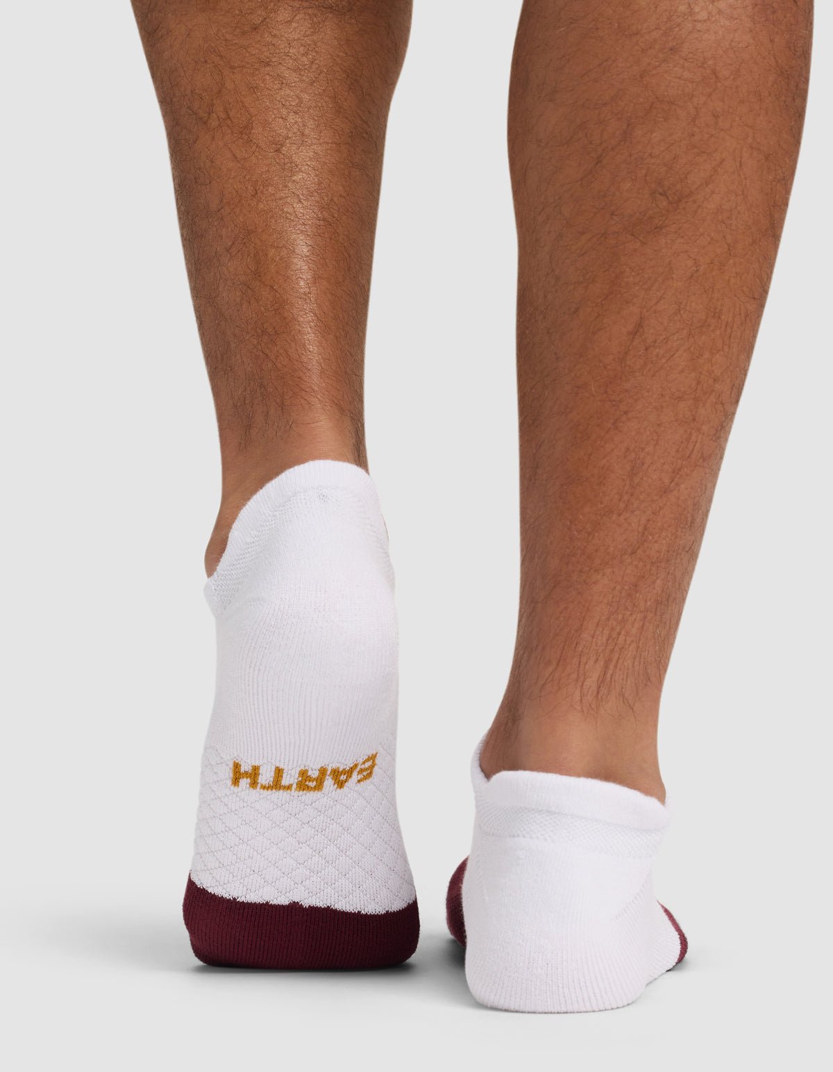 A person wearing Cozy Earth Essential Ankle Socks in white with maroon soles and "EARTH" in yellow on the left heel; the back of their lower legs is shown against a plain background.|Color:Burgundy