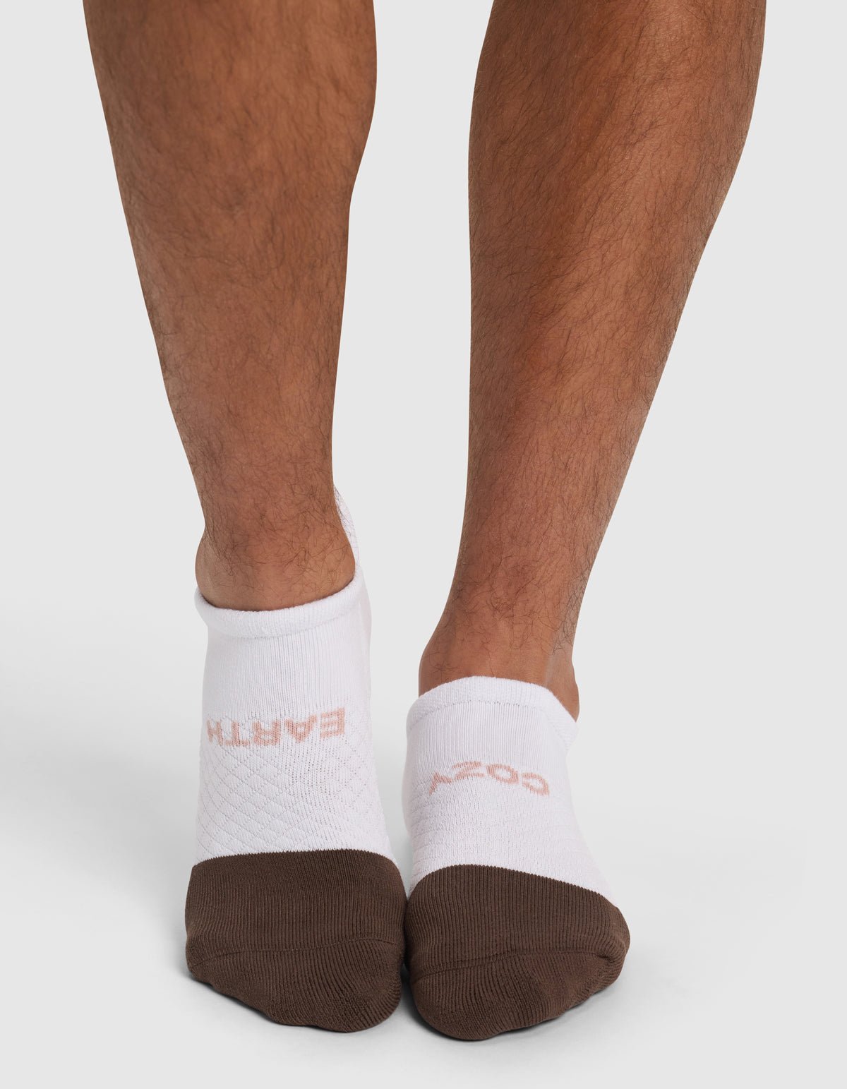 A person wears mismatched white Essential Ankle Socks by Cozy Earth with brown toes; one reads "EARTH" and the other "COZY" in light pink. They stand against a plain white background.|Color:Cedar