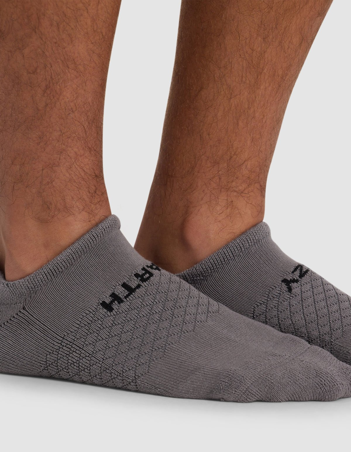 A close-up of a person's lower legs wearing Cozy Earth's Essential Ankle Sock in light gray with a subtle pattern and black text, standing on a light gray surface.|Color:Stone