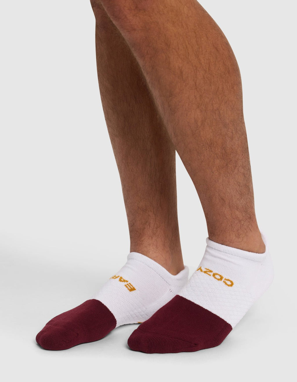 A person wearing Cozy Earth Essential Ankle Socks with maroon toes and yellow text stands on a light surface, showing only their lower legs and feet.|Color:Burgundy