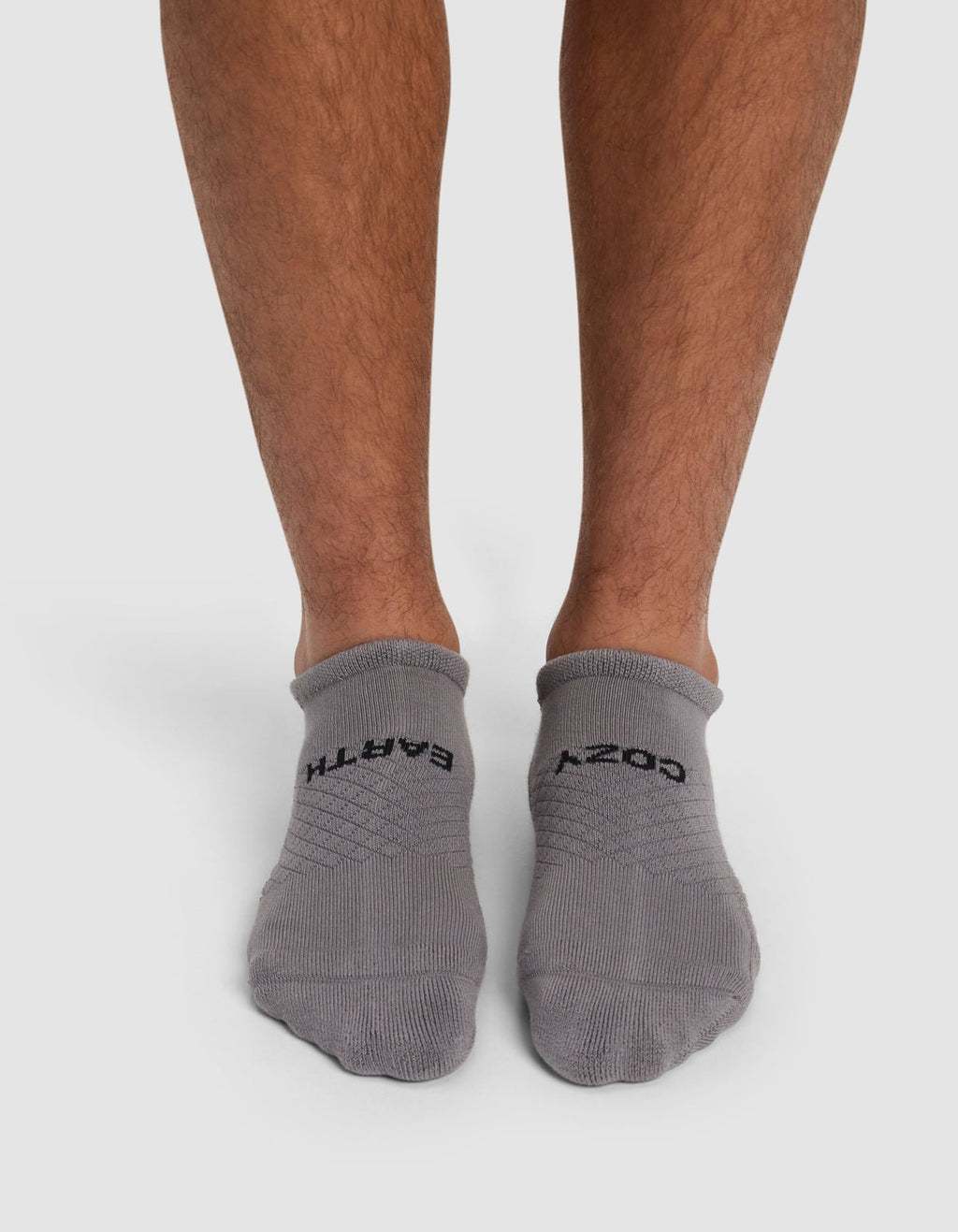 A person, with only their lower legs and feet visible, stands on a white background wearing Cozy Earth Essential Ankle Socks in gray. The left sock reads "EARTH" and the right reads "COZY.|Color:Stone