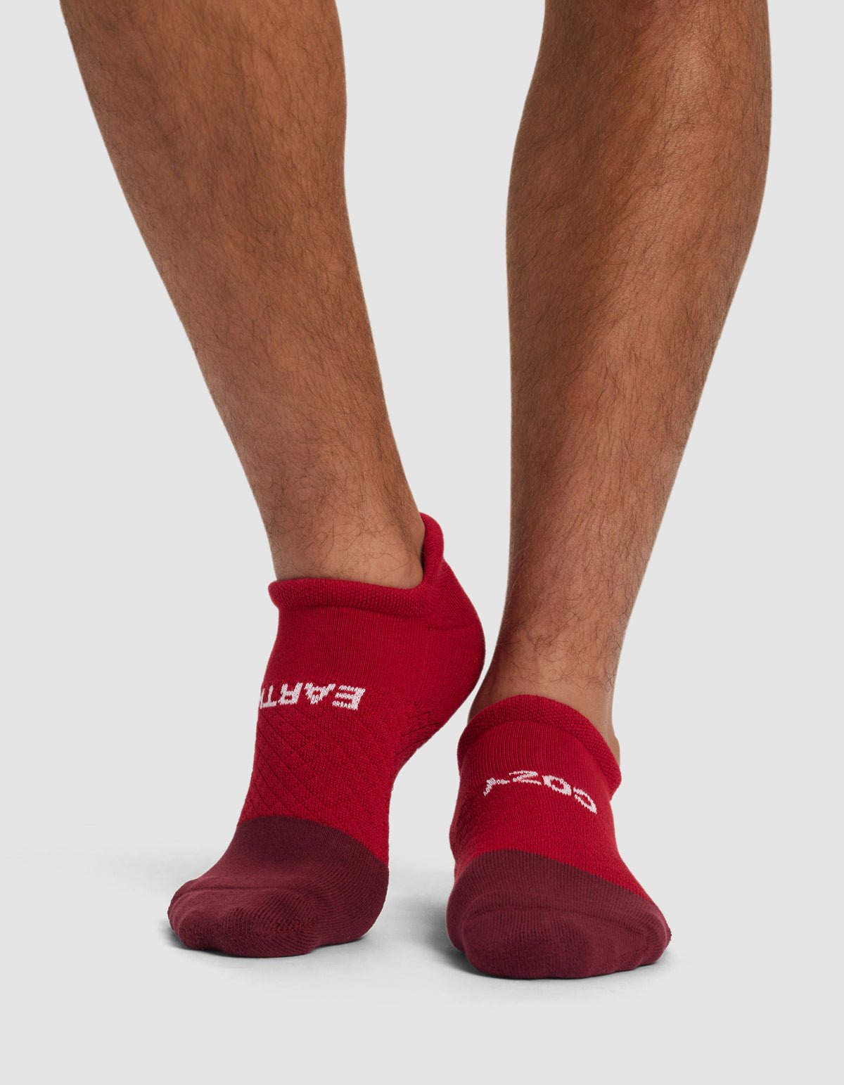 A person wearing Cozy Earth Essential Ankle Socks in red, featuring "EARTH" on the left and "GOOD" on the right at the top, stands on a white surface. Only their lower legs and feet are visible.|Color:Crimson