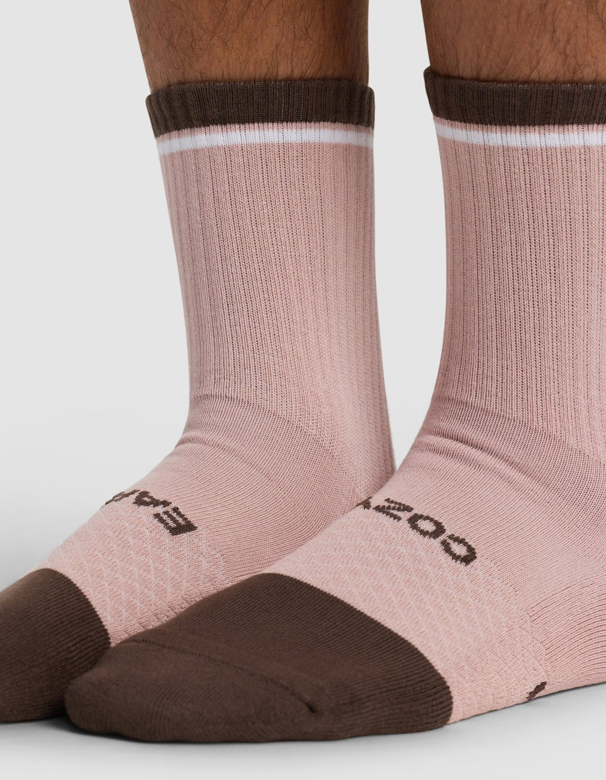 Close-up of a person's lower legs and feet wearing Cozy Earth Essential Calf Socks in light pink with dark brown toes, heels, cuffs, and a thin white stripe near the cuff. The brand name is partially visible on the socks.|Color:Cedar