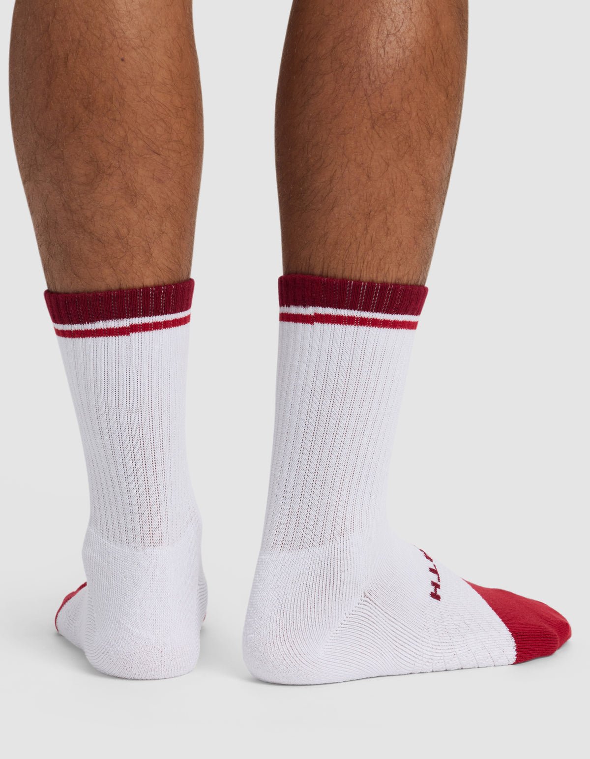 A pair of legs wearing Cozy Earth Essential Calf Socks in white, featuring red toes, heels, and top stripes. These ribbed socks display partially visible text on the soles against a plain white background.|Color:Crimson