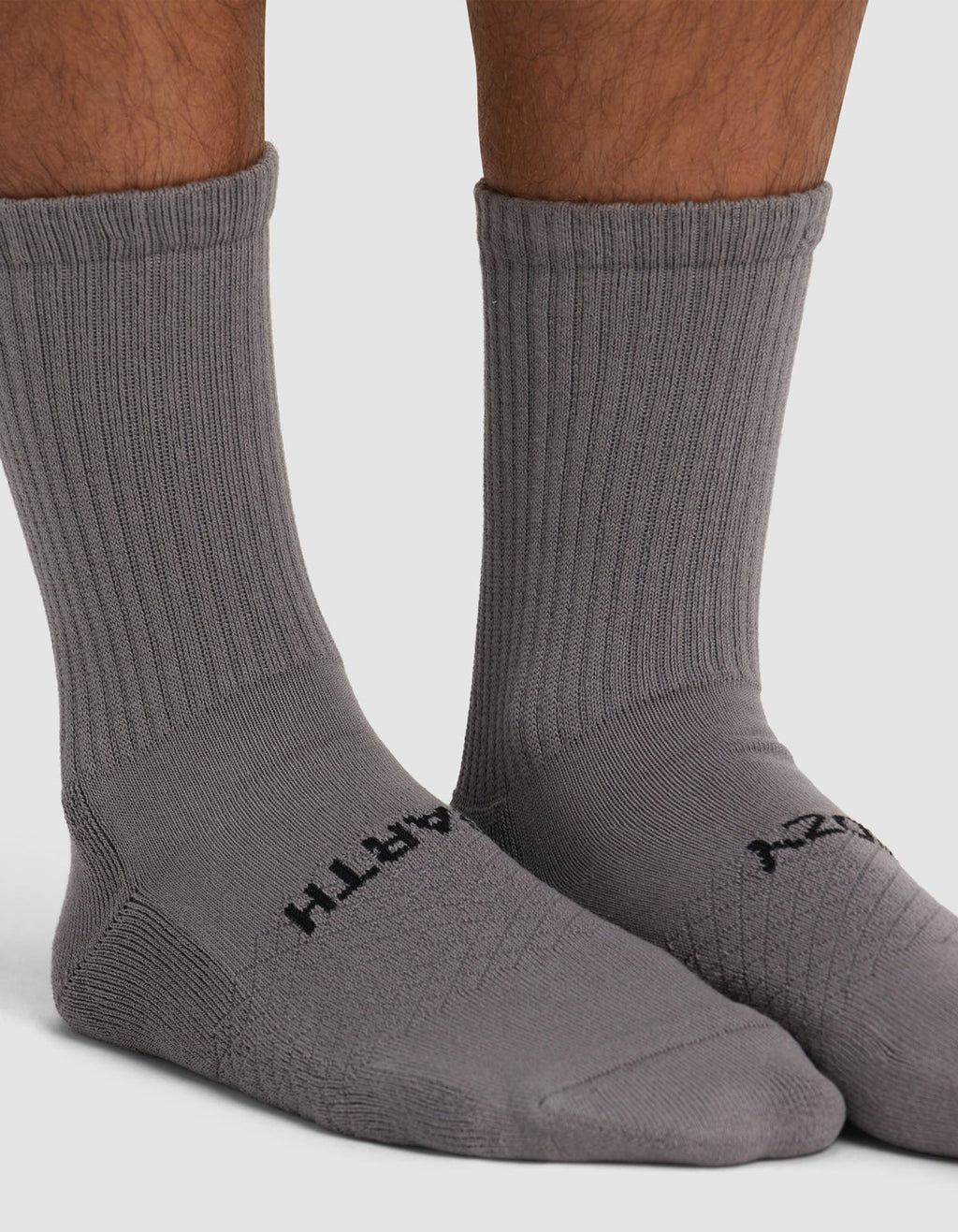 Close-up of a person wearing Cozy Earth Essential Calf Socks in gray with black text near the toes, standing on a white surface. Only the lower legs and feet are visible.|Color:Stone