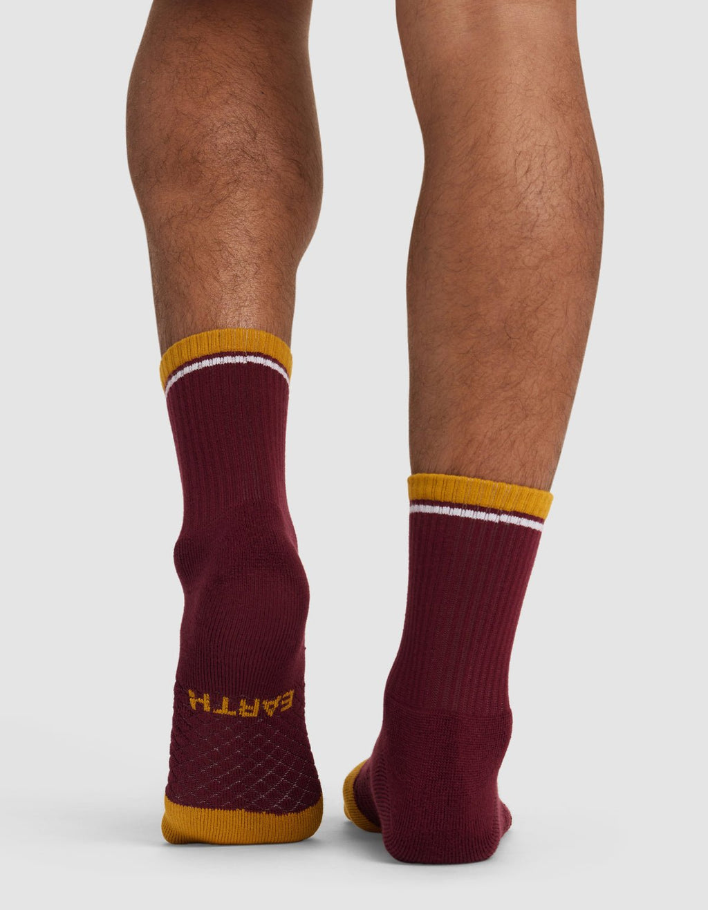 Back view of a person wearing Cozy Earth's Essential Calf Sock—mismatched maroon socks with gold toes and cuffs, white stripes near the top, and "EARTH" on one sole. The person stands on a light background.|Color:Burgundy