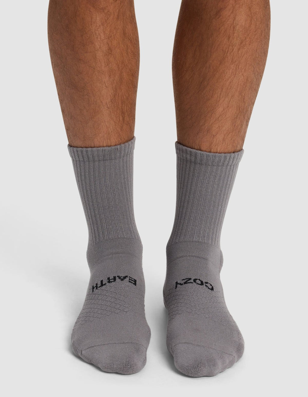 A person stands on a white surface wearing Cozy Earth Essential Calf Socks in gray—the left sock reads "EARTH" and the right sock reads "COZY" in black text.|Color:Stone