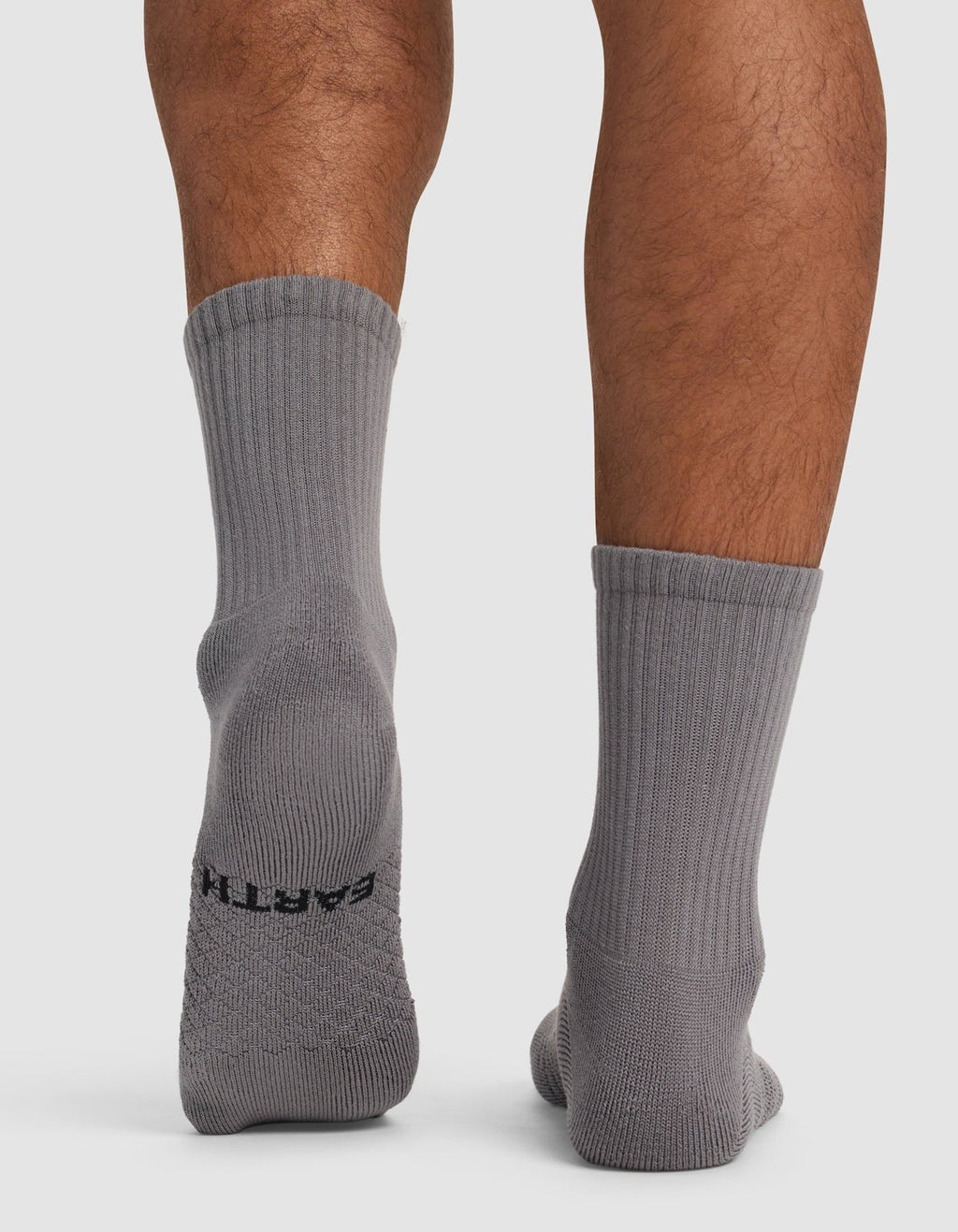 A person stands on a white surface wearing Cozy Earth Essential Calf Socks in gray. The back of the socks and lower legs are visible, with "EARTH" in black text on the sole of the left sock.|Color:Stone