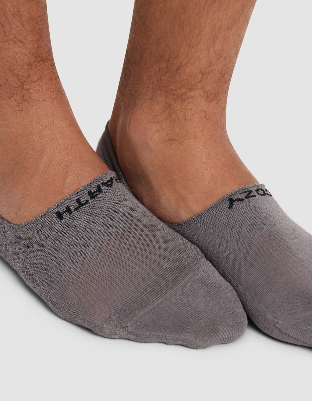 Close-up of a person's feet wearing Cozy Earth Essential No-Show Socks in gray with black text near the cuffs, standing on a white background.|Color:Stone