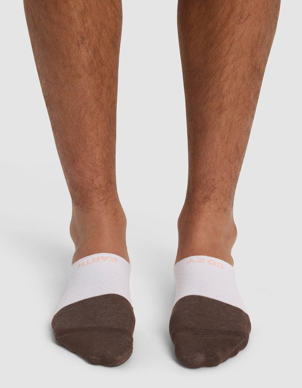 Close-up of a person's lower legs and feet wearing Cozy Earth's Essential No-Show Sock in white and brown, standing on a plain white background.|Color:Cedar