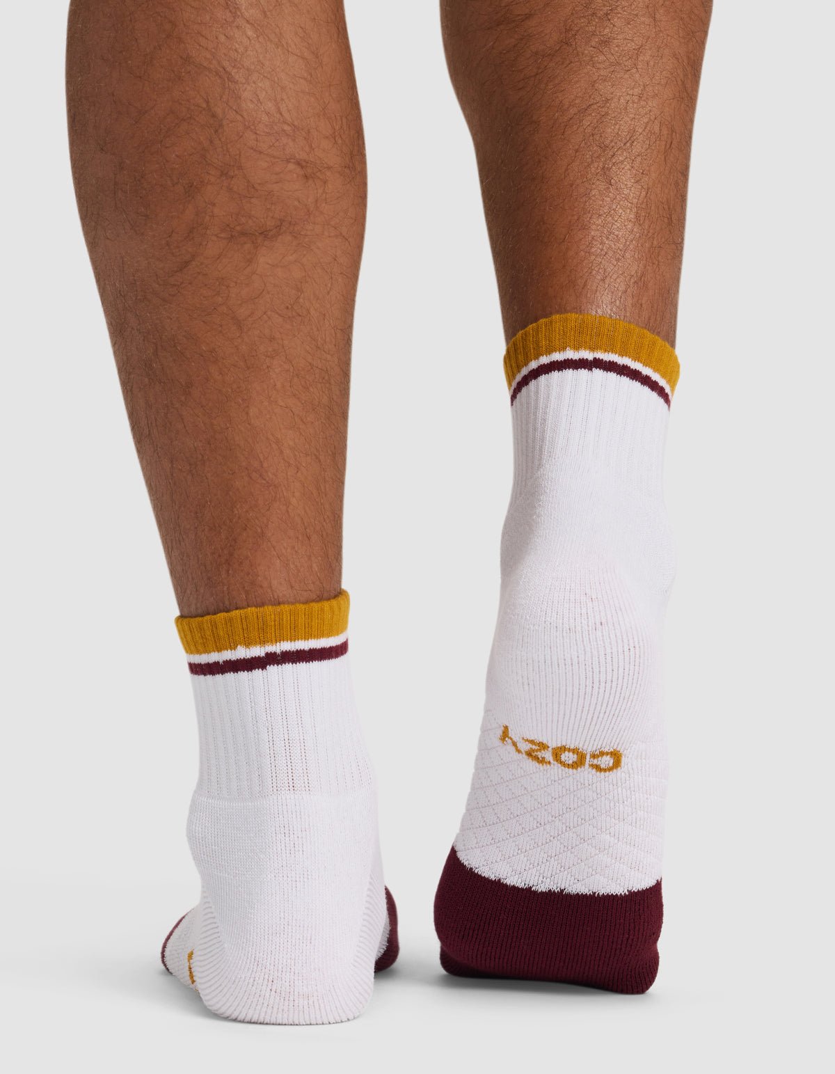 A close-up of the back of lower legs wearing Cozy Earth Essential Quarter Socks—white ankle socks with maroon and gold striped cuffs, maroon heels, and "COZY" in gold lettering on one sole.|Color:Burgundy