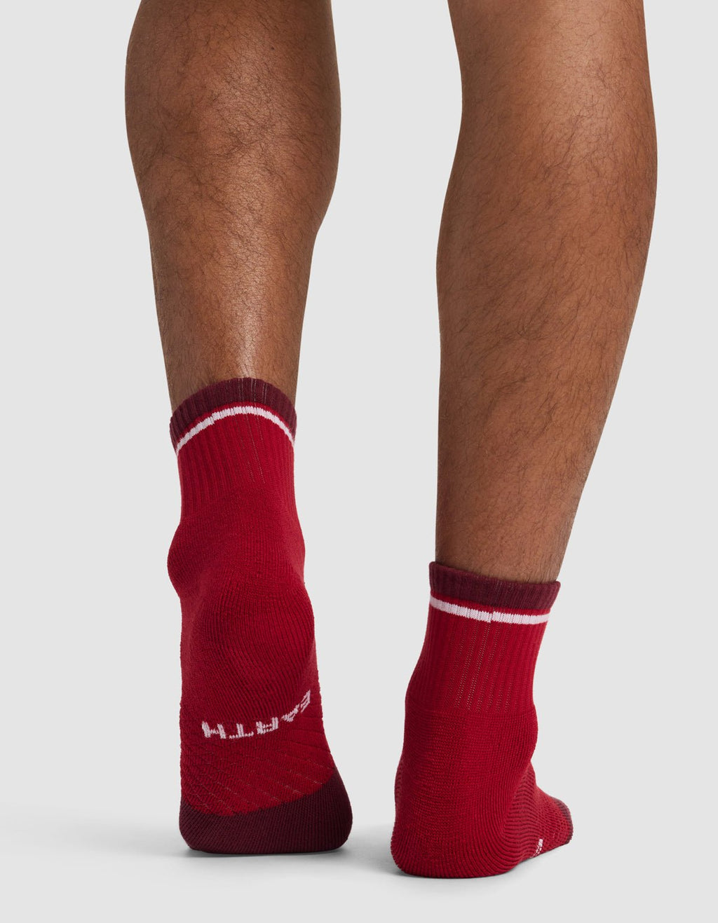 Someone stands on a light-colored surface, showing the back of their legs in Cozy Earth Essential Quarter Socks—red with white stripes. The word "EARTH" is visible on the heel of the left sock.|Color:Crimson