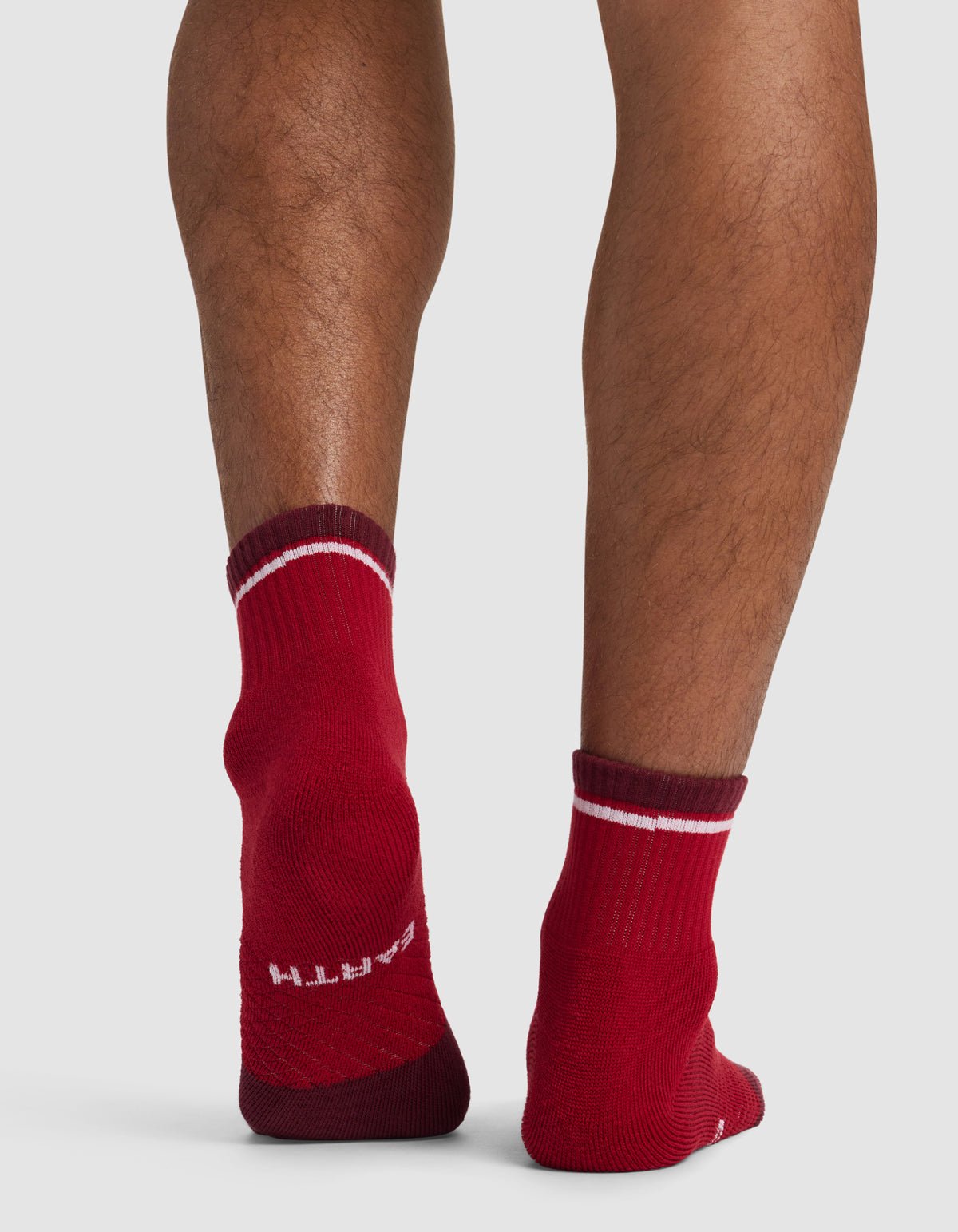 Someone stands on a light-colored surface, showing the back of their legs in Cozy Earth Essential Quarter Socks—red with white stripes. The word "EARTH" is visible on the heel of the left sock.|Color:Crimson