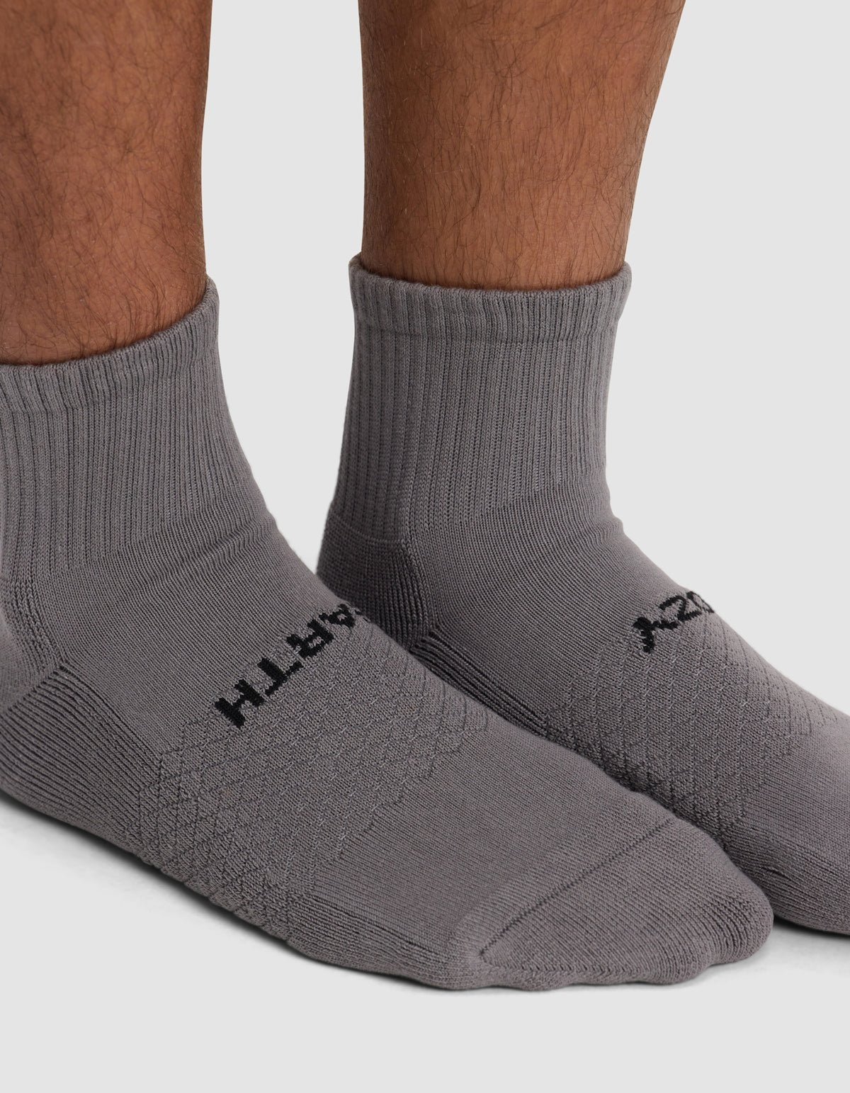 Close-up of a person wearing Cozy Earth's Essential Quarter Sock in gray with black text, standing on a white background.|Color:Stone
